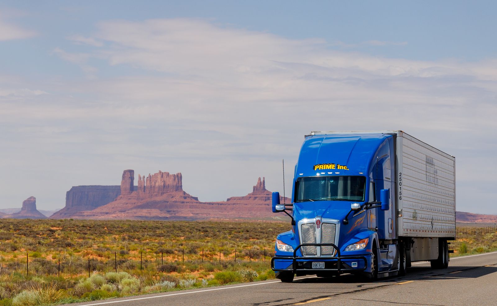 Blue Kenworth semi truck driving a US desert highway past Monument Valley.